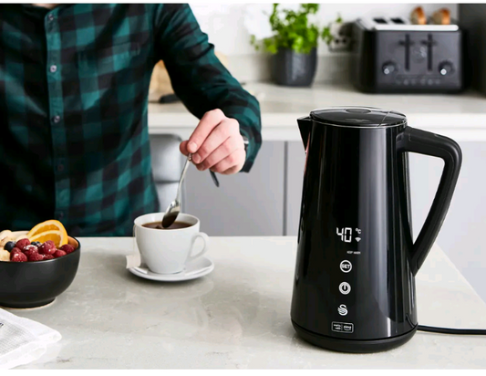 Person stirring coffee with a black electric kettle on a kitchen counter.